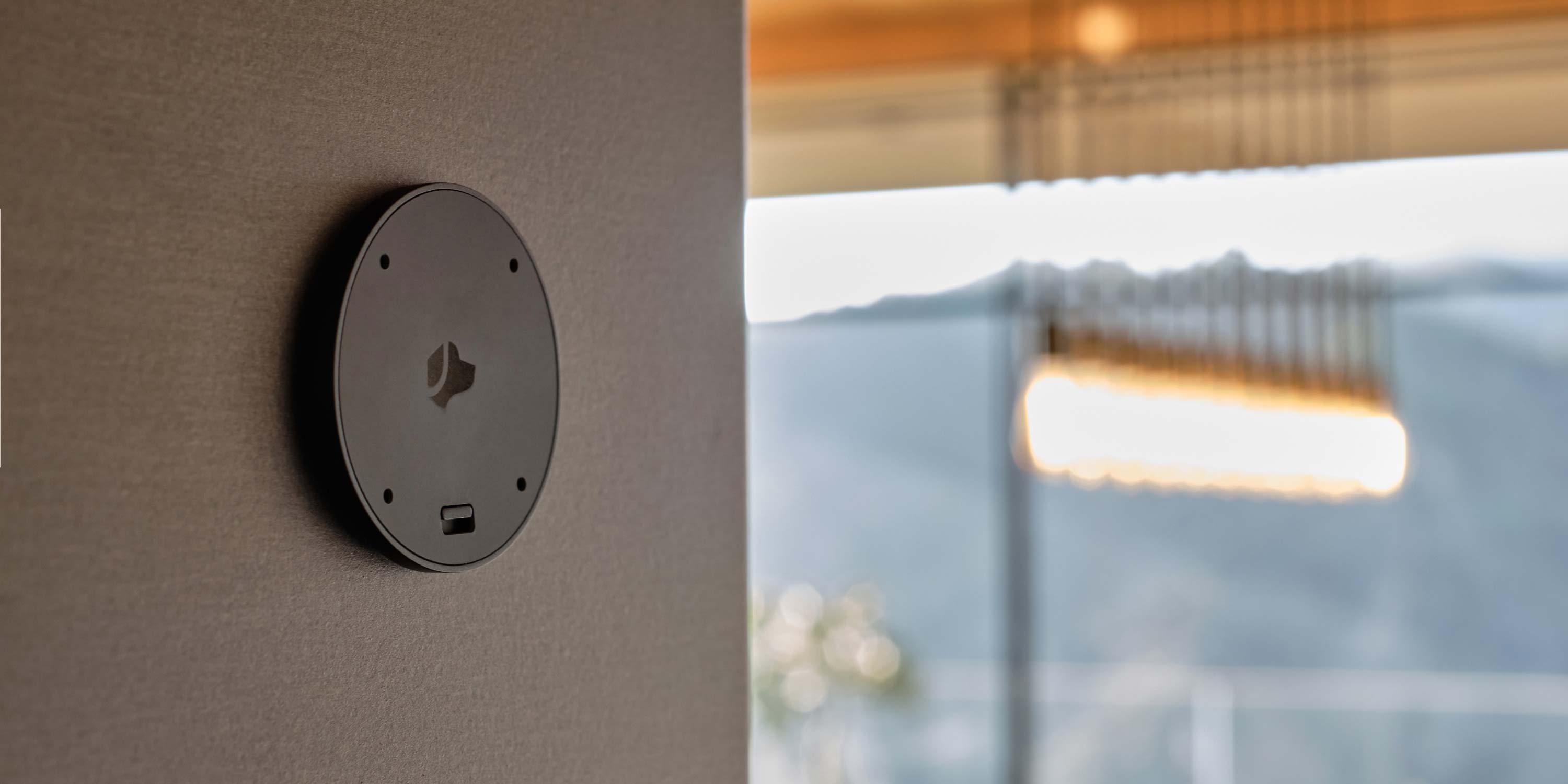 A Josh AI wall-mounted circular device with a dog logo, placed on a textured wall in a modern indoor setting with a blurred background of a well-lit room.