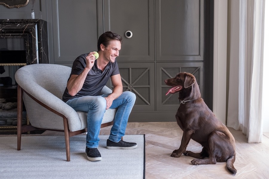 A man plays fetch with his dog in the living room — a Josh.ai voice control device is on the wall.